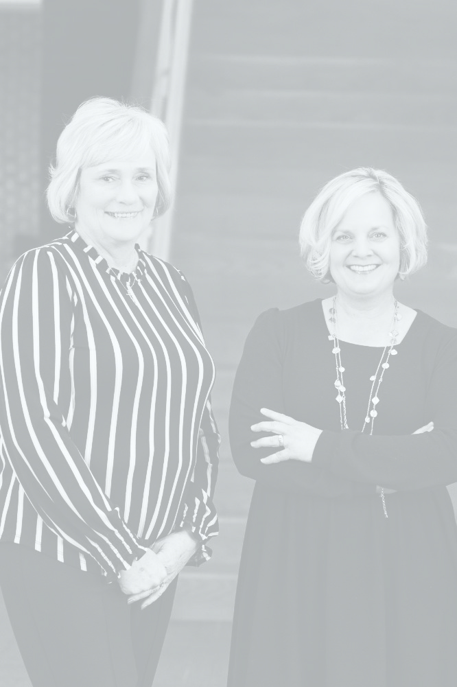 Group portrait of three professional women smiling in business attire, two with blonde hair and one with brunette hair, standing in an indoor setting with a blurred background.